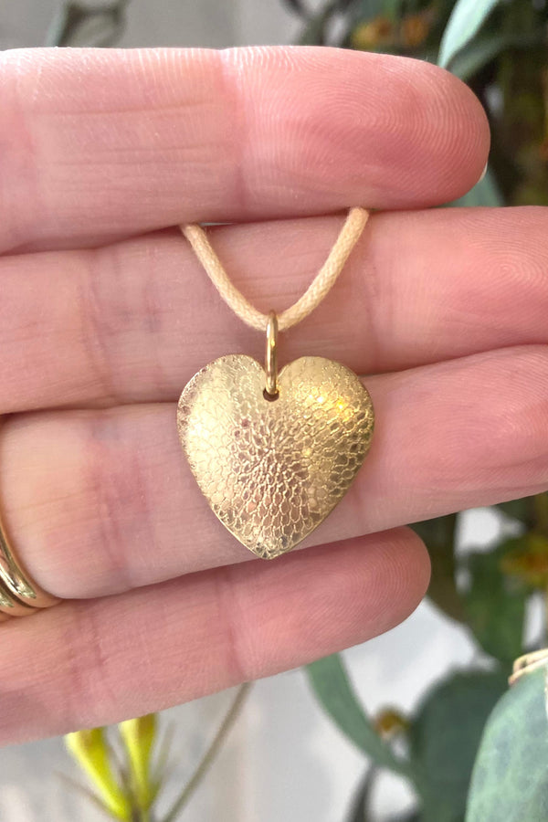 Gold heart-shaped engraved pendant with a detailed chrysanthemum petal design, strung on a cotton cord, held in the hand that made it. 