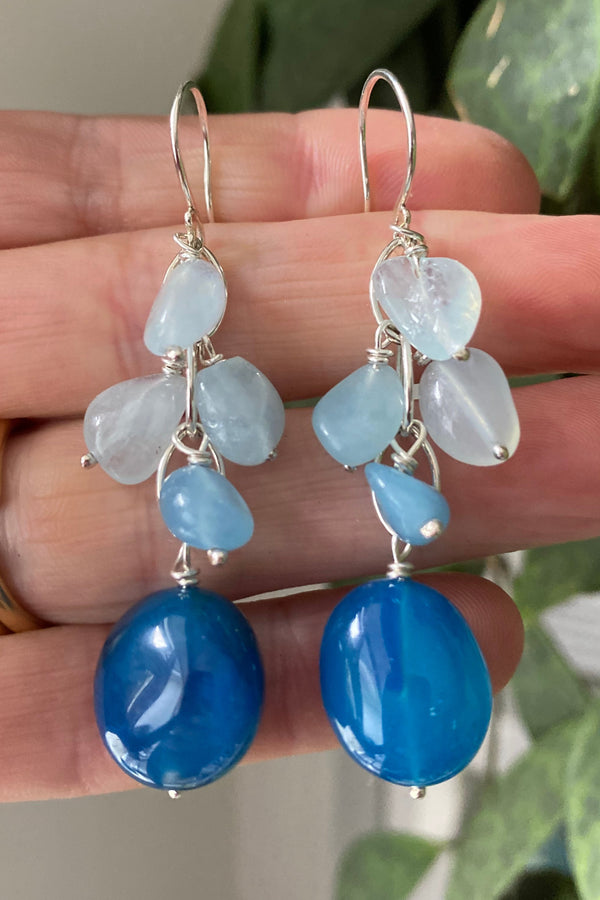 Chalcedony and aquamarine gemstone waterfall earrings in shades of dark and light blue, held by the hand that made them. 