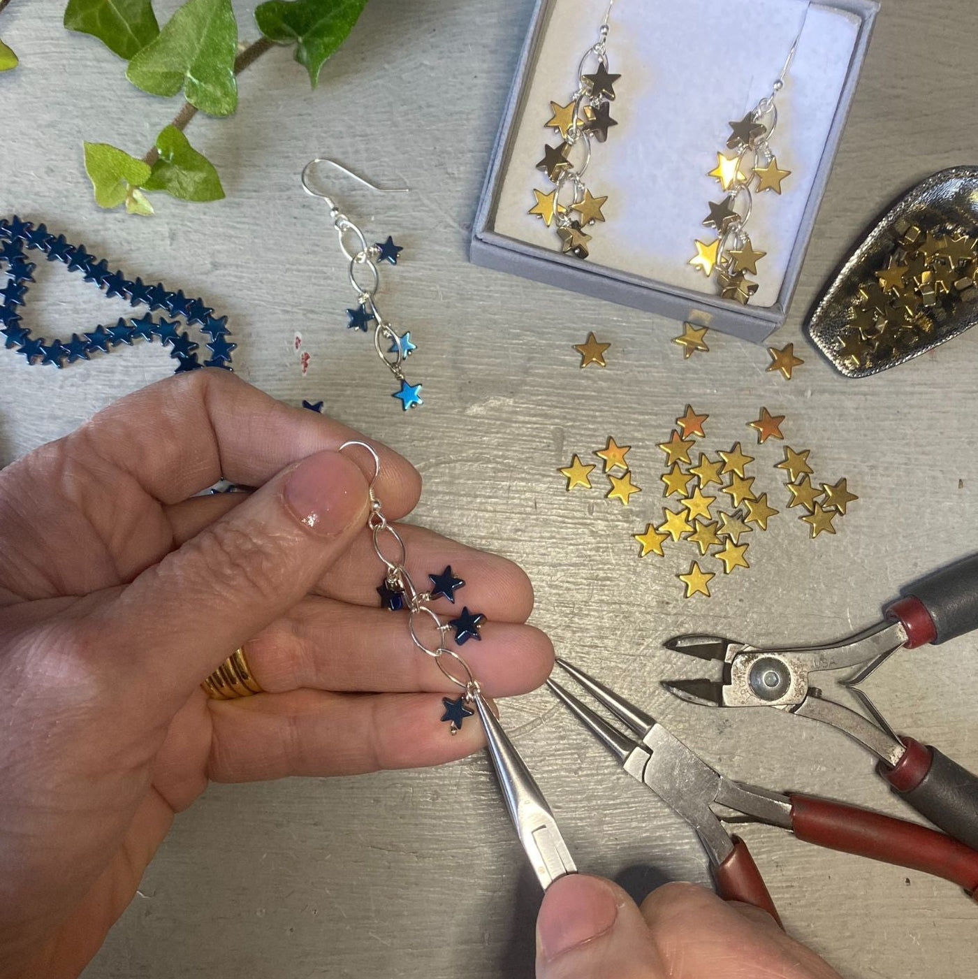 Hands crafting star-shaped dangle earrings with silver and blue beads using metal pliers on a light gray tabletop, with a box of finished earrings and loose star embellishments nearby.