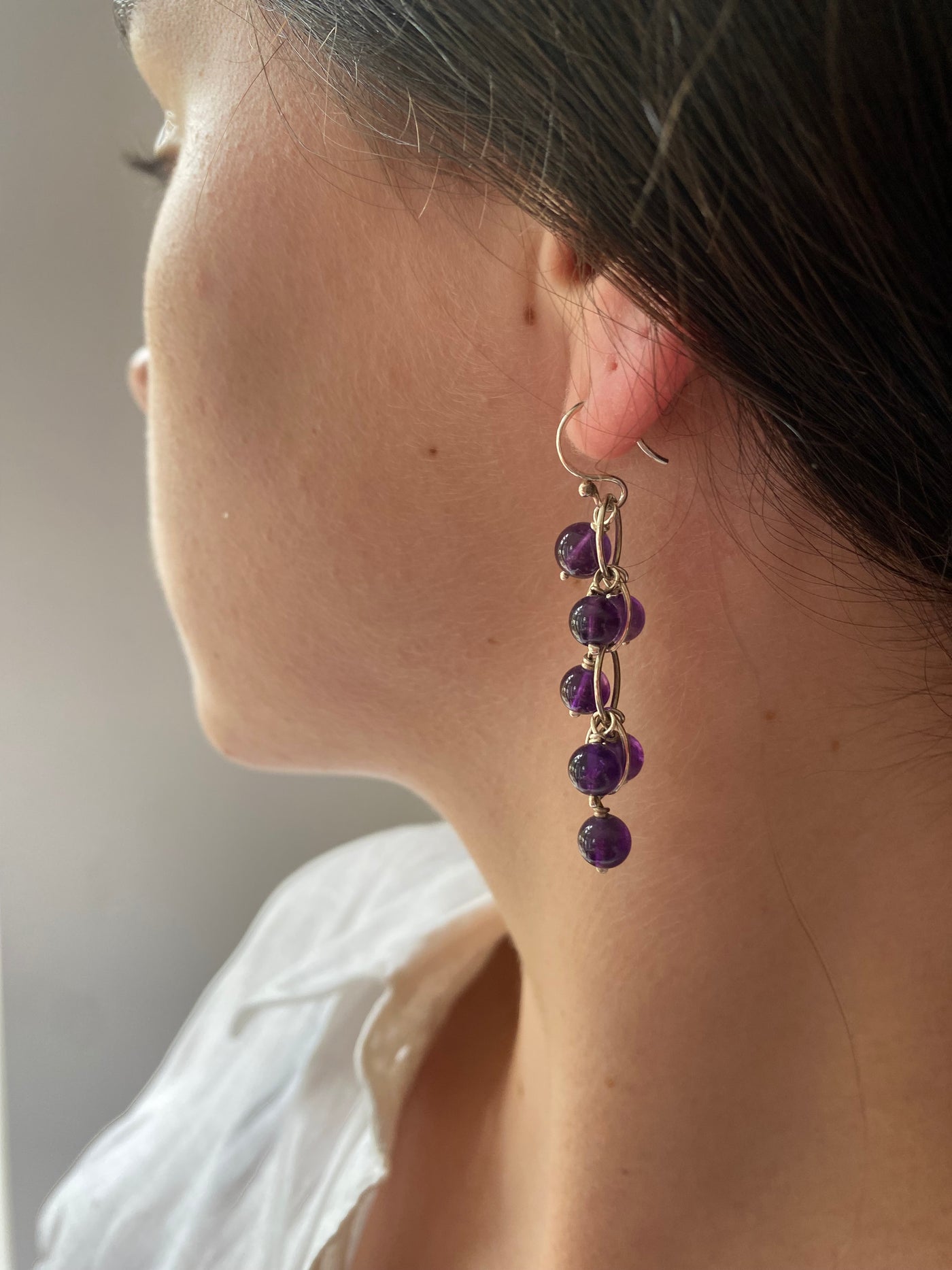 Close-up of a waterfall earring with purple  amethyst gemstone beads worn by a person in a white blouse. 