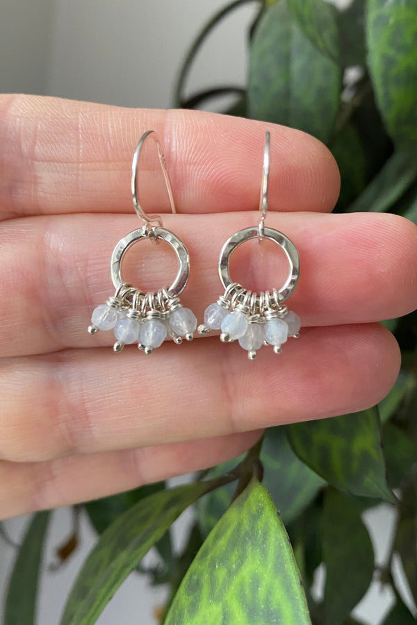 Silver earrings with small aquamarine stones held between fingers against a green leafy background