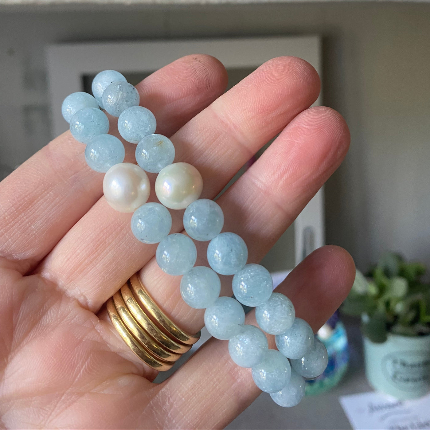 A light blue aquamarine gemstone beaded bracelet held on the hand that made it, with a blurred indoor background. 