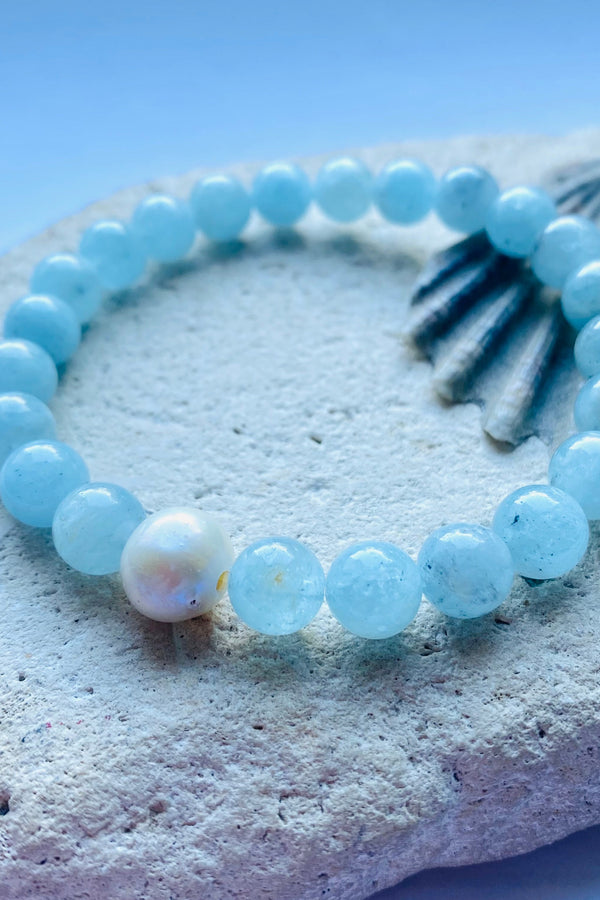 Pale blue beaded aquamarine gemstone bracelet with a freshwater pearl accent on a rock and shell background
