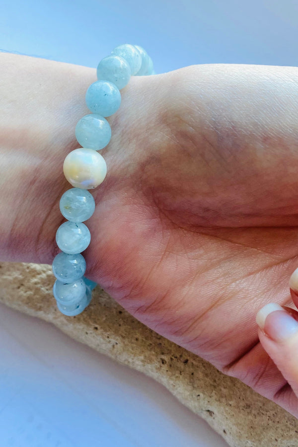 Hand wearing a pale blue aquamarine gemstone bracelet with light blue beads and a freshwater pearl accent, resting on a stone surface on a light background