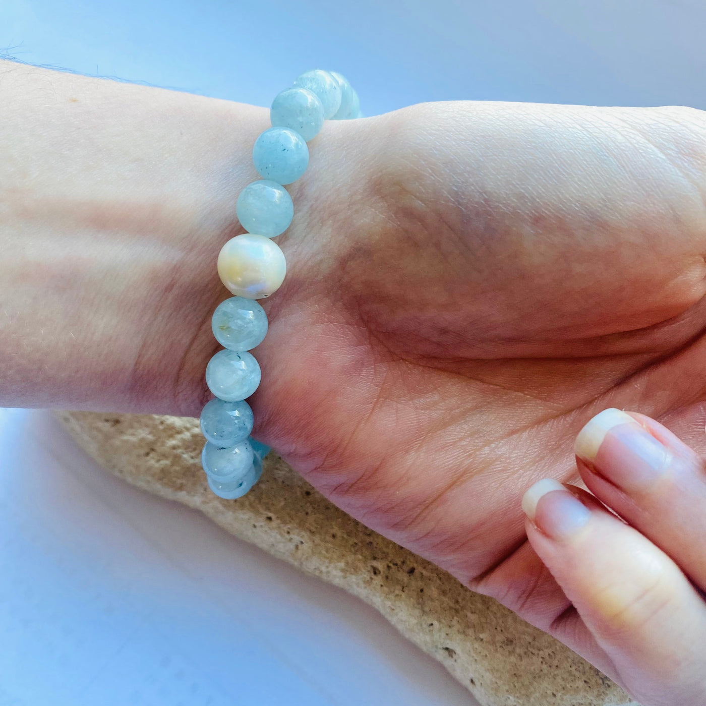 Hand wearing a pale blue aquamarine gemstone bracelet with light blue beads and a freshwater pearl accent, resting on a stone surface on a light background