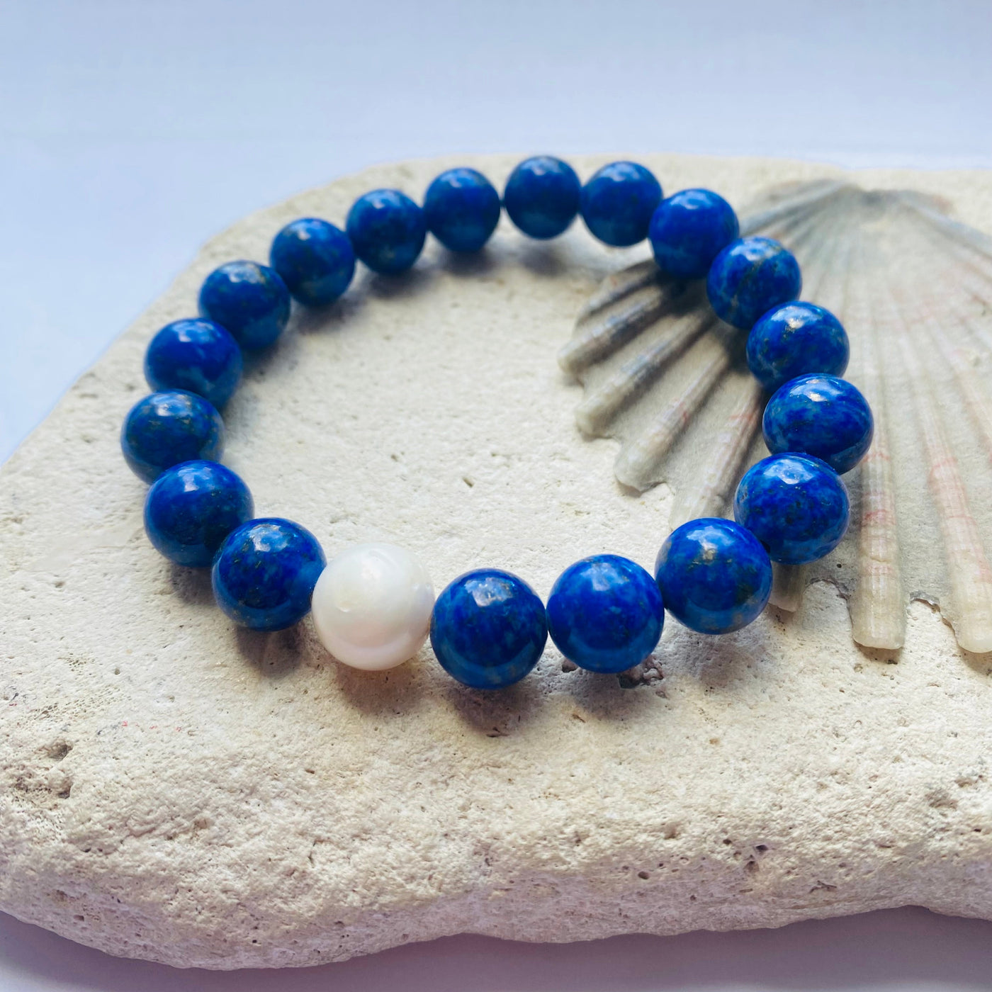 Blue beaded lapis lazuil bracelet with a white freshwater pearl on a rock with a shell background. 