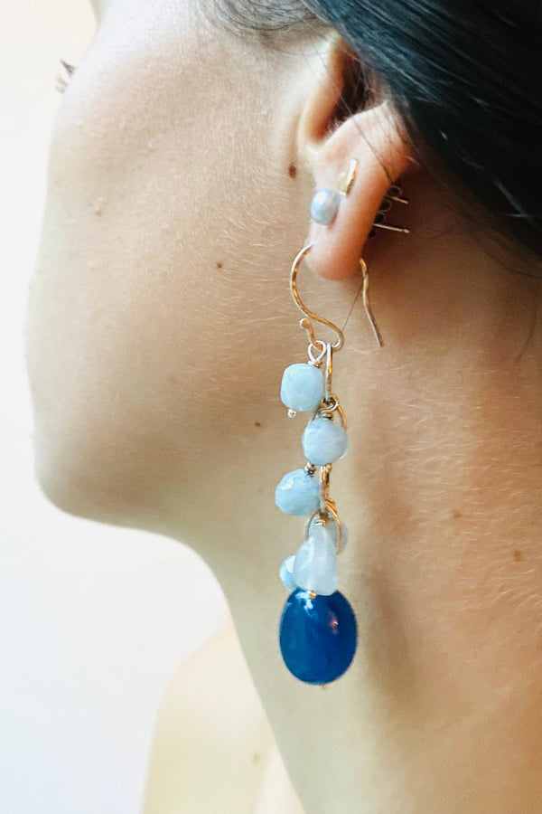 Close-up of a person wearing an earring with deep blue chalcedony and pale blue aquamarine gemstones on a light background