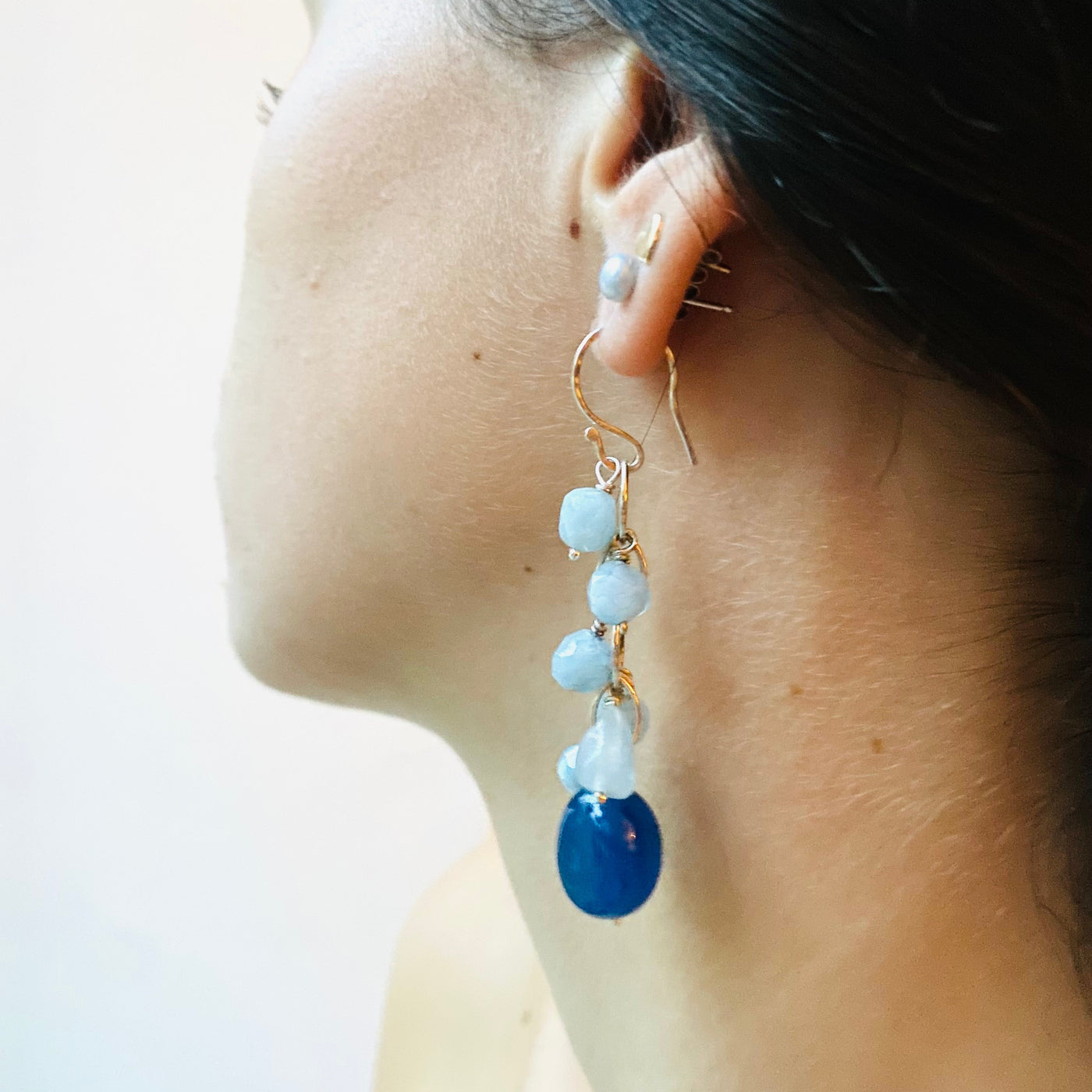 Close-up of a person wearing an earring with deep blue chalcedony and pale blue aquamarine gemstones on a light background