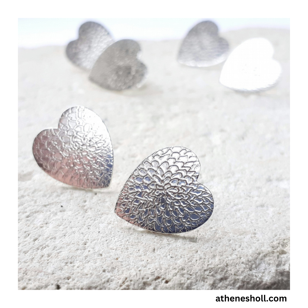 Silver heart-shaped earrings on a textured white surface with 'athenesholl.com' branding.
