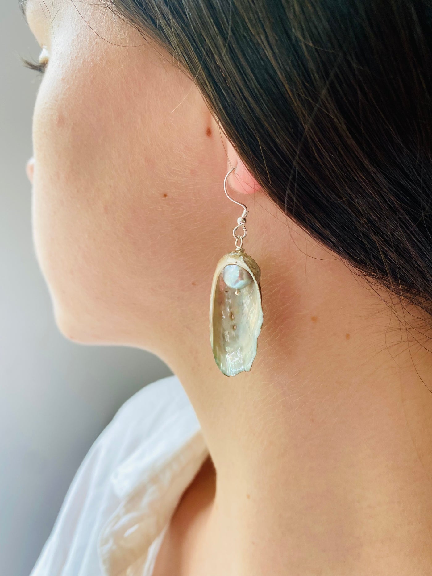Close-up of a Guernsey ormer shell earring worn on a model, earrings has a grey freshwater pearl and an irridescent surface. 