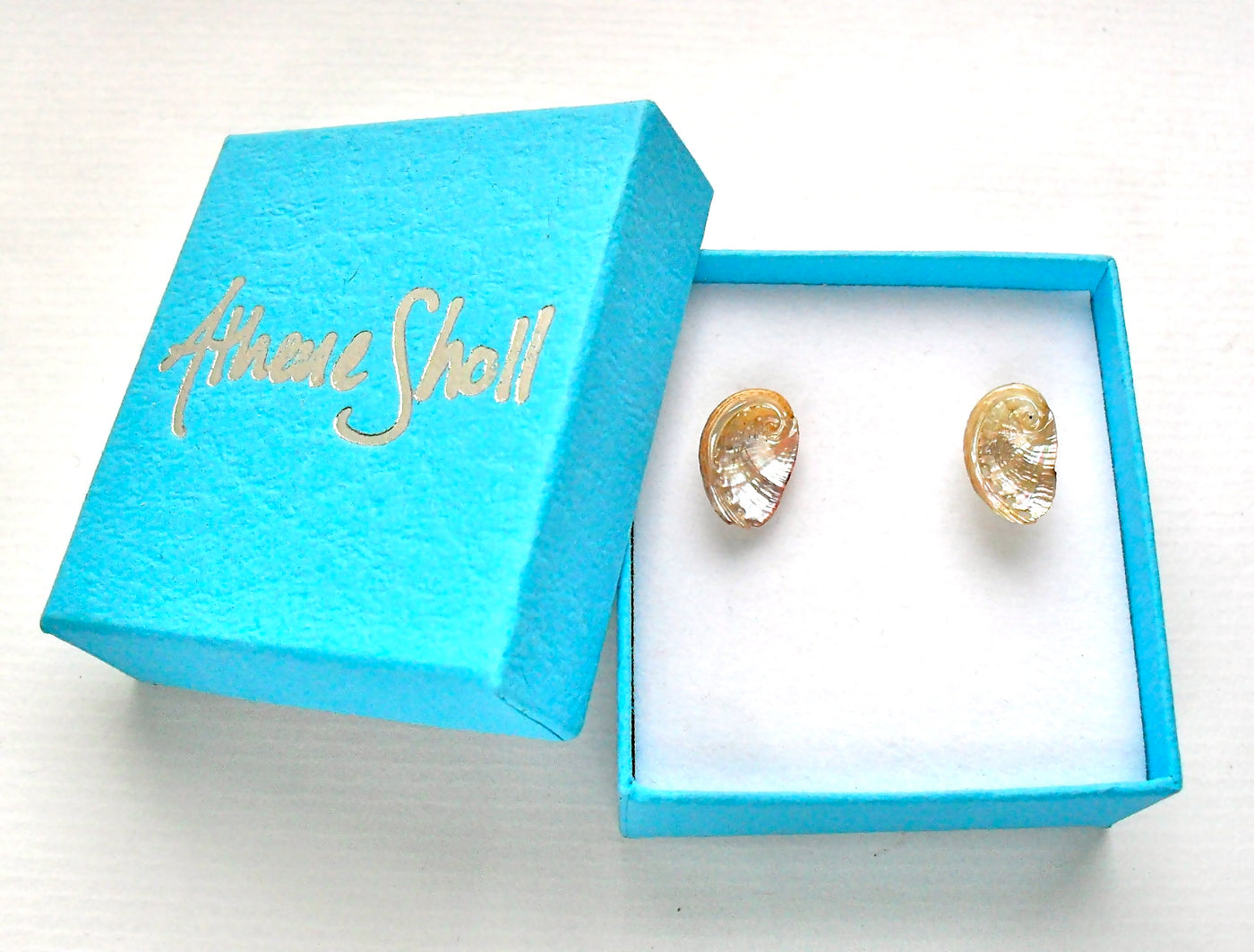 Gold coloured ormer shell earrings in a blue box with 'Athene Sholl' branding.