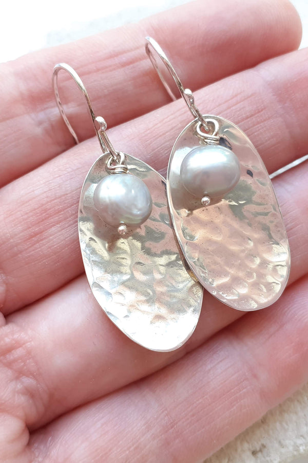 Silver potato peel pie earrings with freshwater pearls held in the hand that made them against a light background