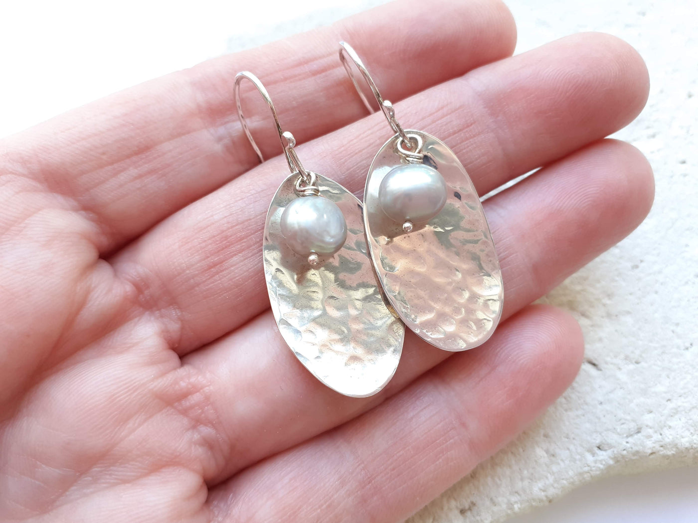 Silver potato peel pie earrings with freshwater pearls held in the hand that made them against a light background