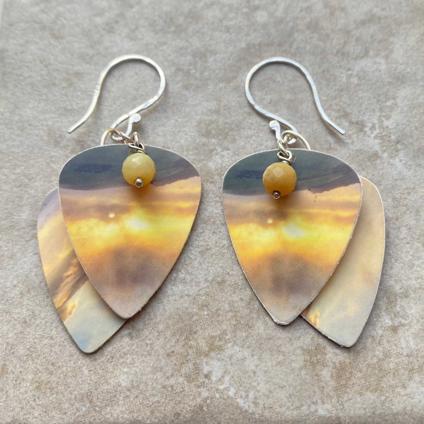 Pair of plectrum shaped earrings with layered metallic yellow and grey sunset design and central bead on a textured surface