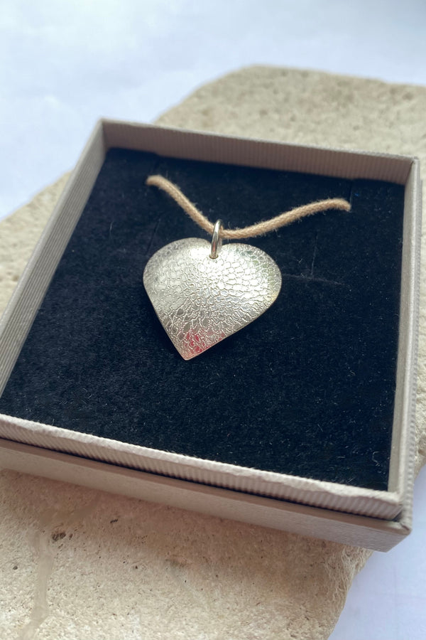 Hand engraved flower design heart-shaped silver pendant in a box on a textured surface