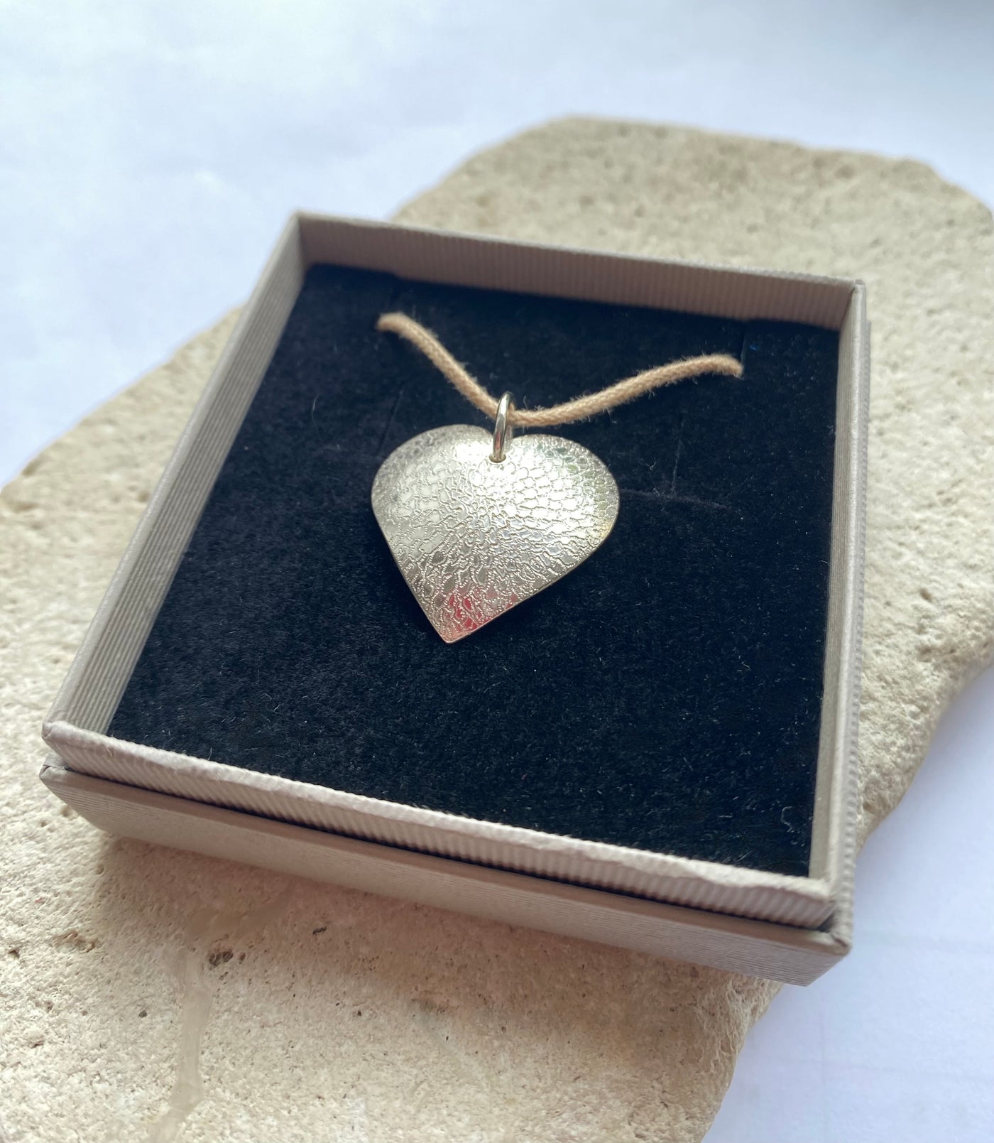 Hand engraved flower design heart-shaped silver pendant in a box on a textured surface