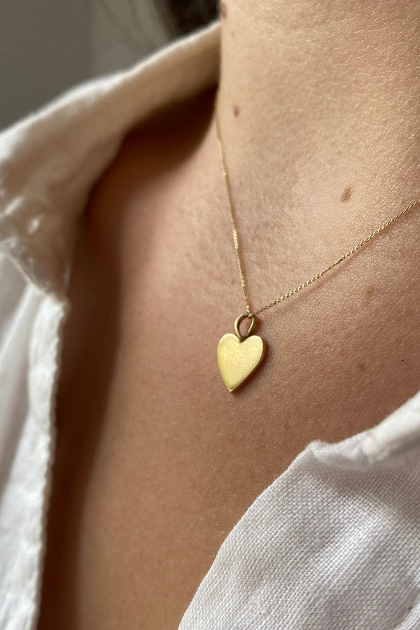 Close-up of a person wearing a white shirt  and a heart-shaped  9ct gold pendant necklace on a neutral background