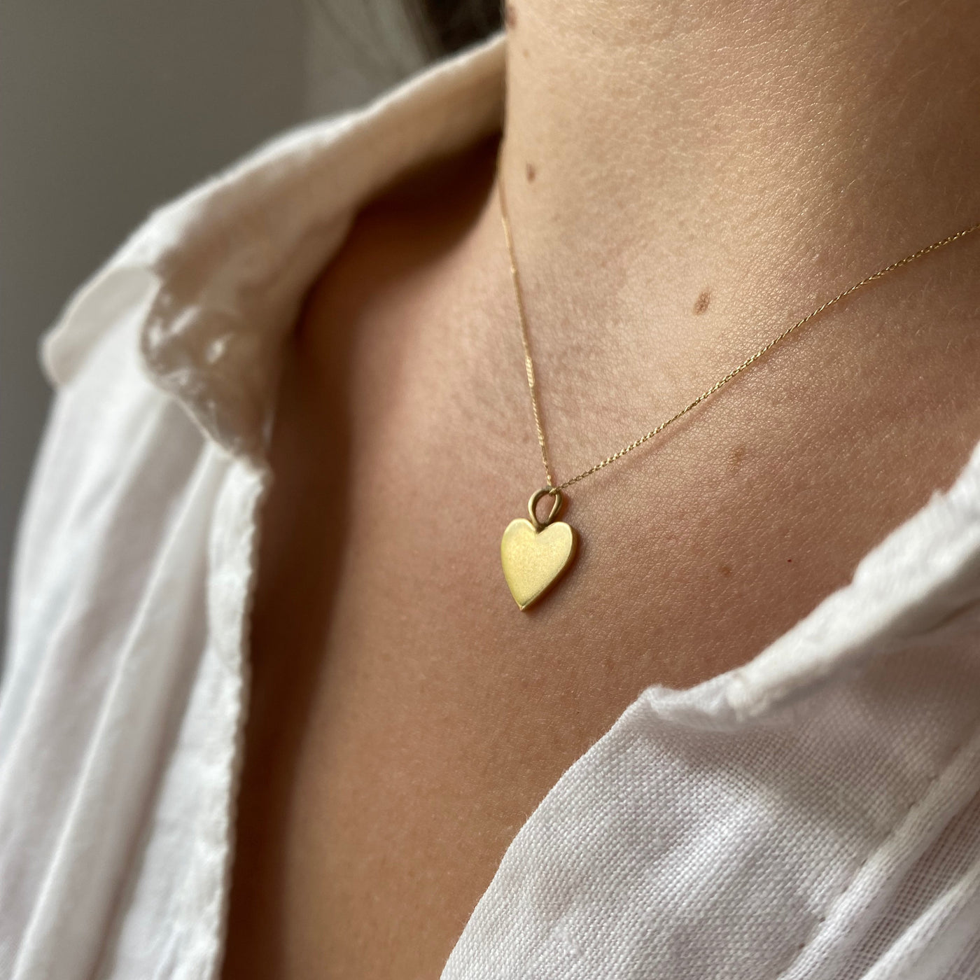 Close-up of a person wearing a white shirt  and a heart-shaped  9ct gold pendant necklace on a neutral background