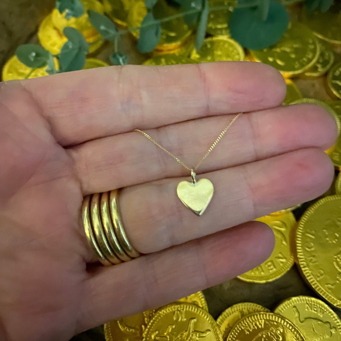  A gold heart-shaped pendant necklace with gold coins and greenery in the background, held on the hand that made it