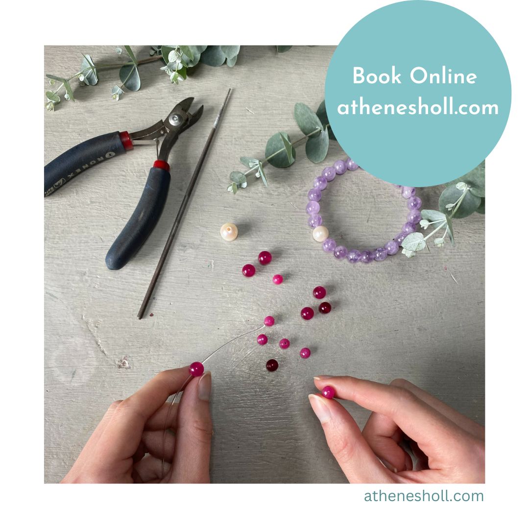 A top-down view of hands beading with pink and red round beads on a gray wooden surface, with jewelry-making tools, eucalyptus leaves, and a purple beaded bracelet nearby. A teal circular badge reads 'Book Online athenesholl.