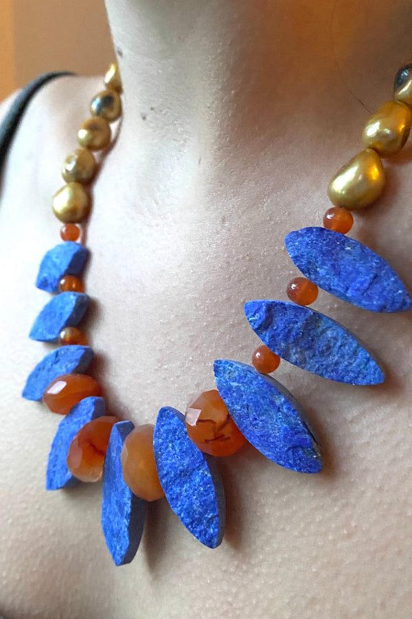 Model wearing a necklace in blue lapis marquise shaped gemstones, with vivid orange carnelia and dyed yellow freshwater pearls. 
