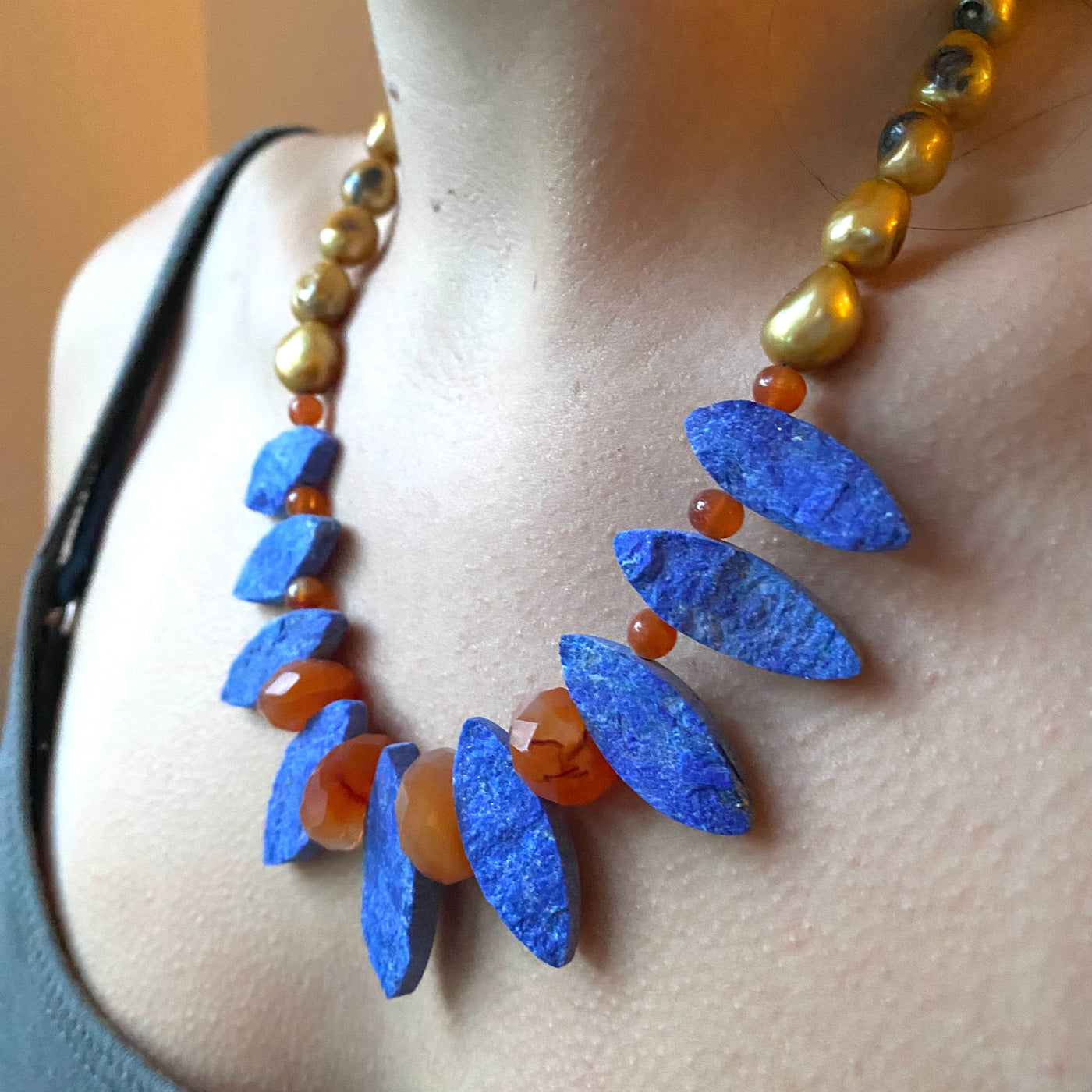 Model wearing a necklace in blue lapis marquise shaped gemstones, with vivid orange carnelia and dyed yellow freshwater pearls. 