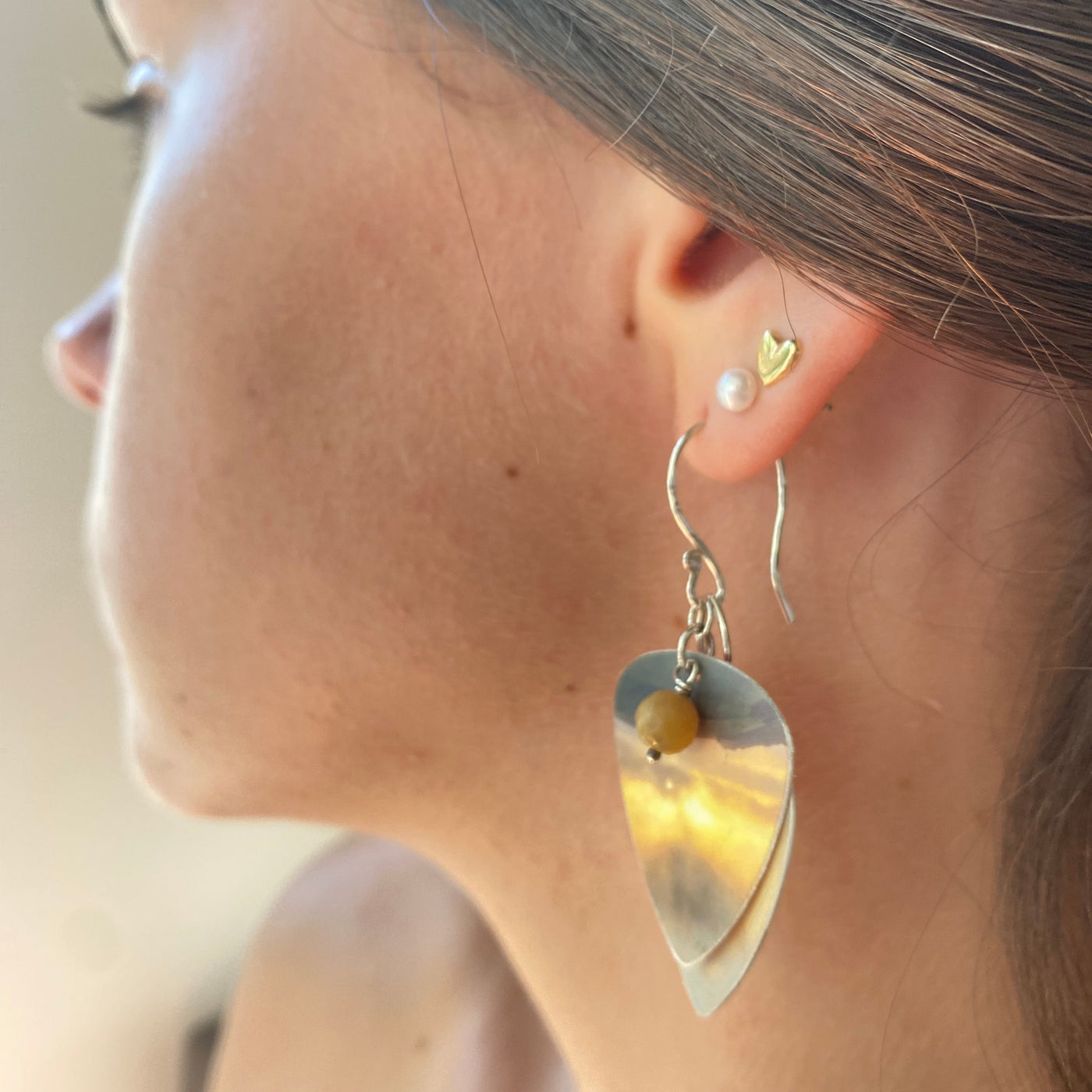 Close-up of a model wearing a silver earring with a heart-shaped pendant, cut from recycled plastic with a yellow semi precious stone drop attached 