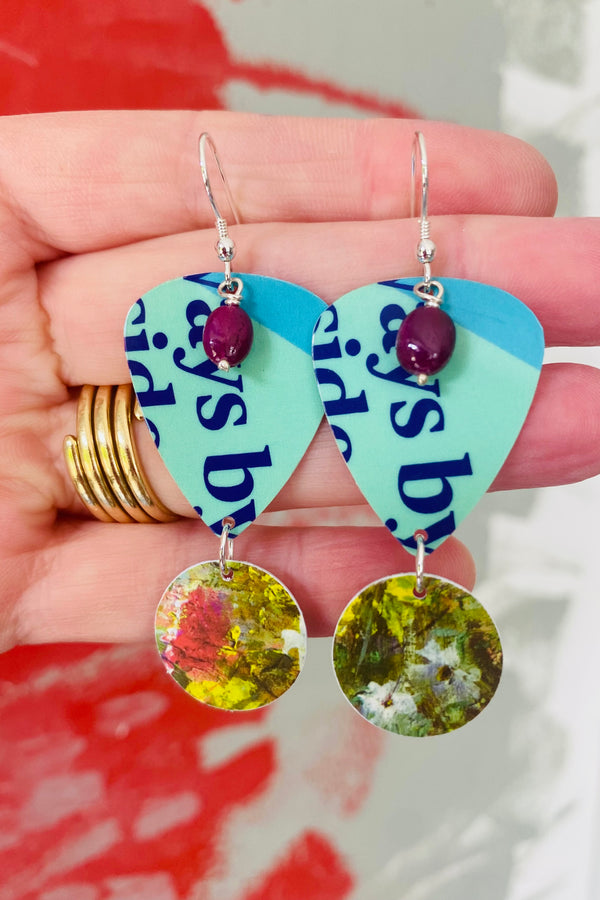 Guitar pick earrings with floral designs and ruby gemstones held by the hand that made them against a blurred background