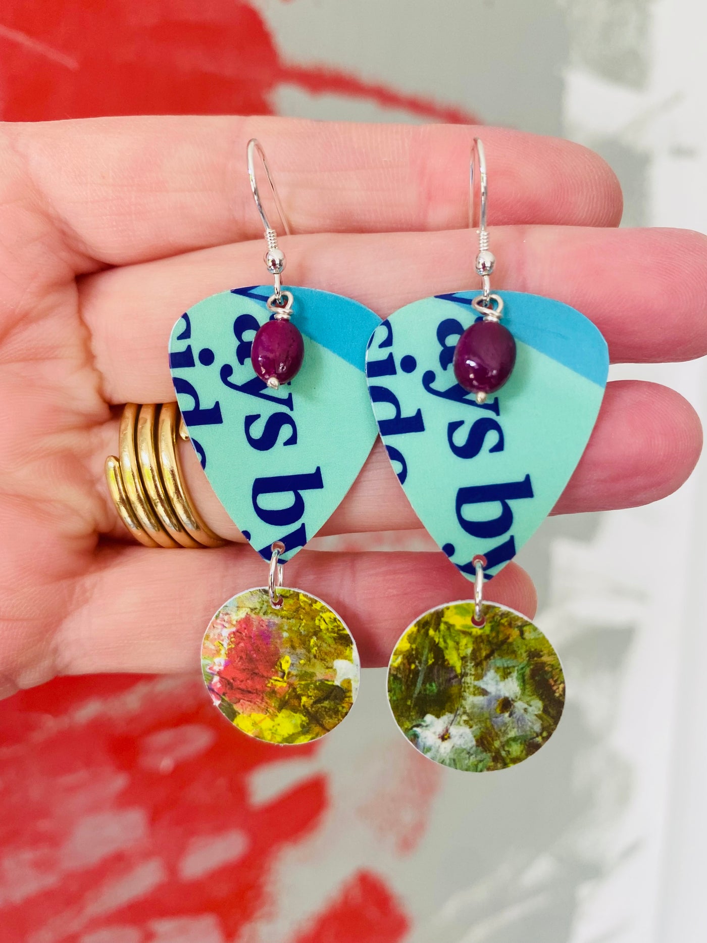 Guitar pick earrings with floral designs and ruby gemstones held by the hand that made them against a blurred background