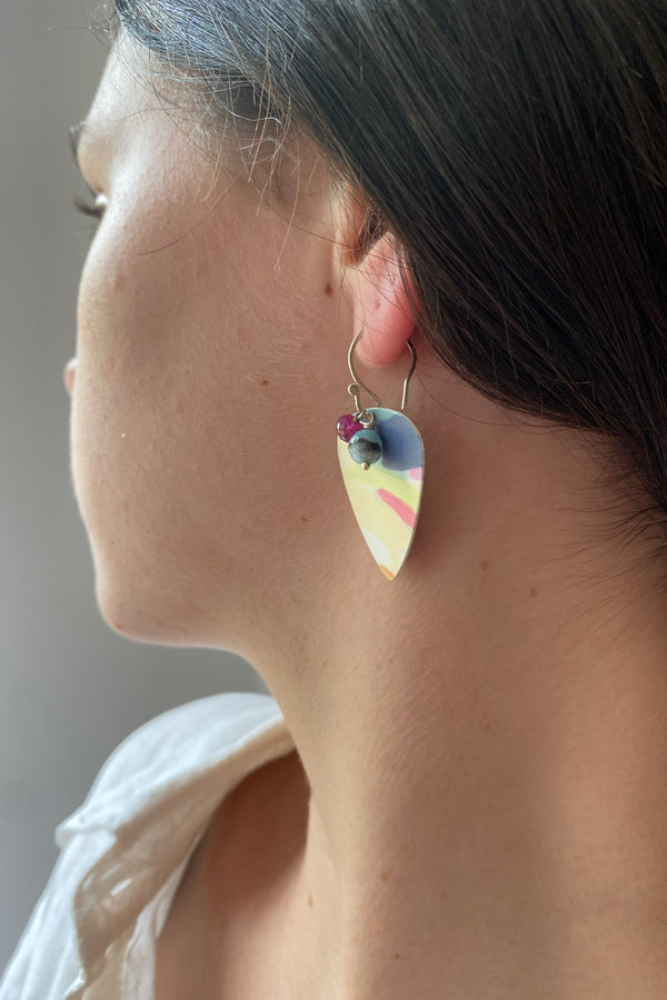 Model waering plectrum shaped earring, with a painterly design of muted colours, and to gemstones, amazonite and ruby, as accents. 