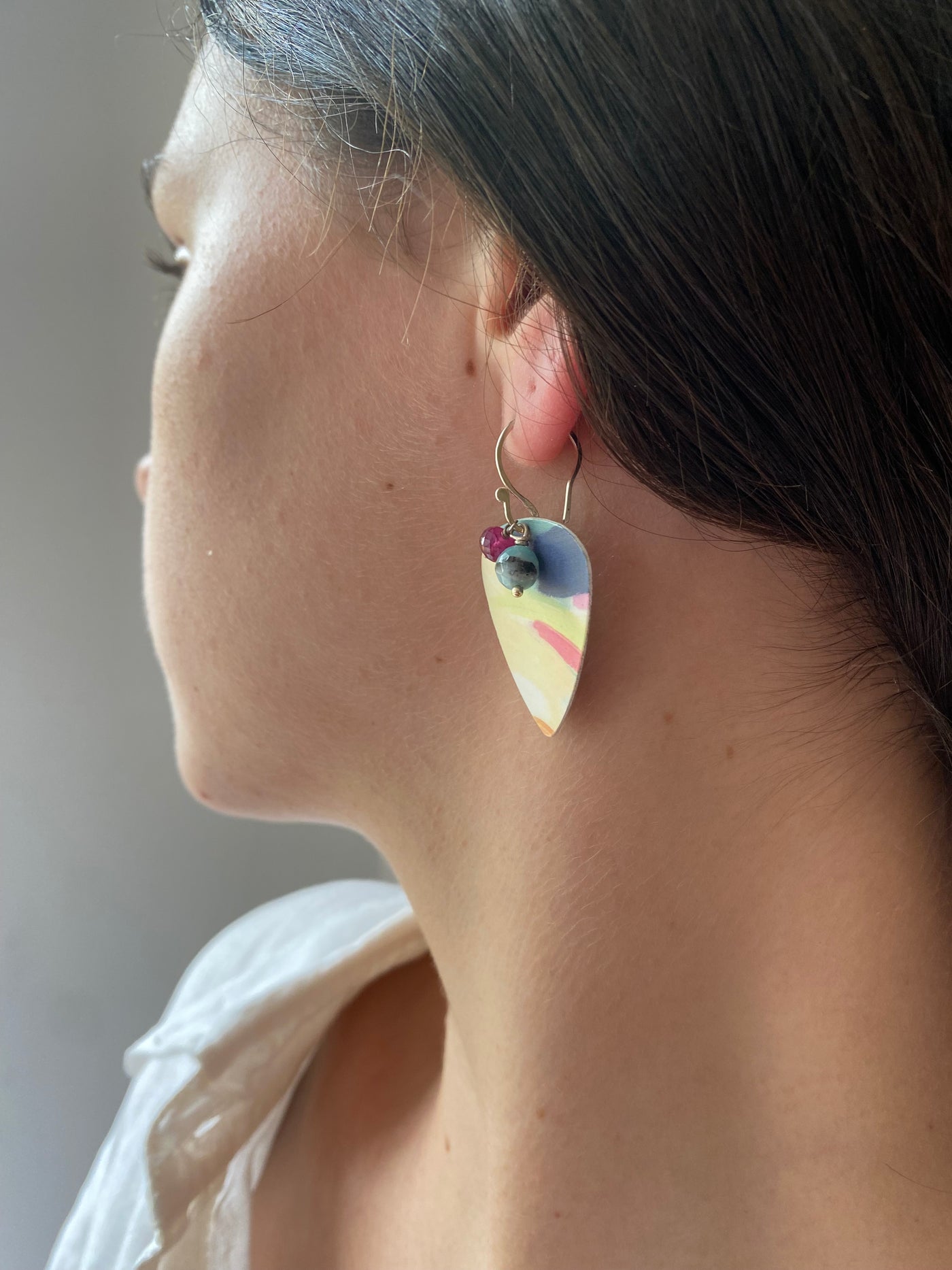 Model waering plectrum shaped earring, with a painterly design of muted colours, and to gemstones, amazonite and ruby, as accents. 