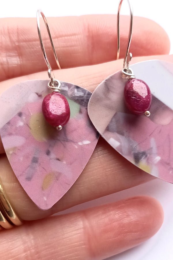 Pink floral guitar pick earrings with a central ruby gemstone bead held in the hand that made them on a white background