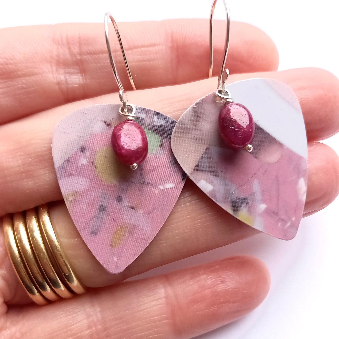 Pink floral guitar pick earrings with a central ruby gemstone bead held in the hand that made them on a white background