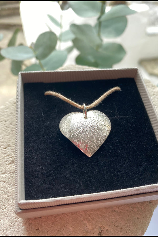Silver heart-shaped pendant on a cotton cord in a box with greenery in the background