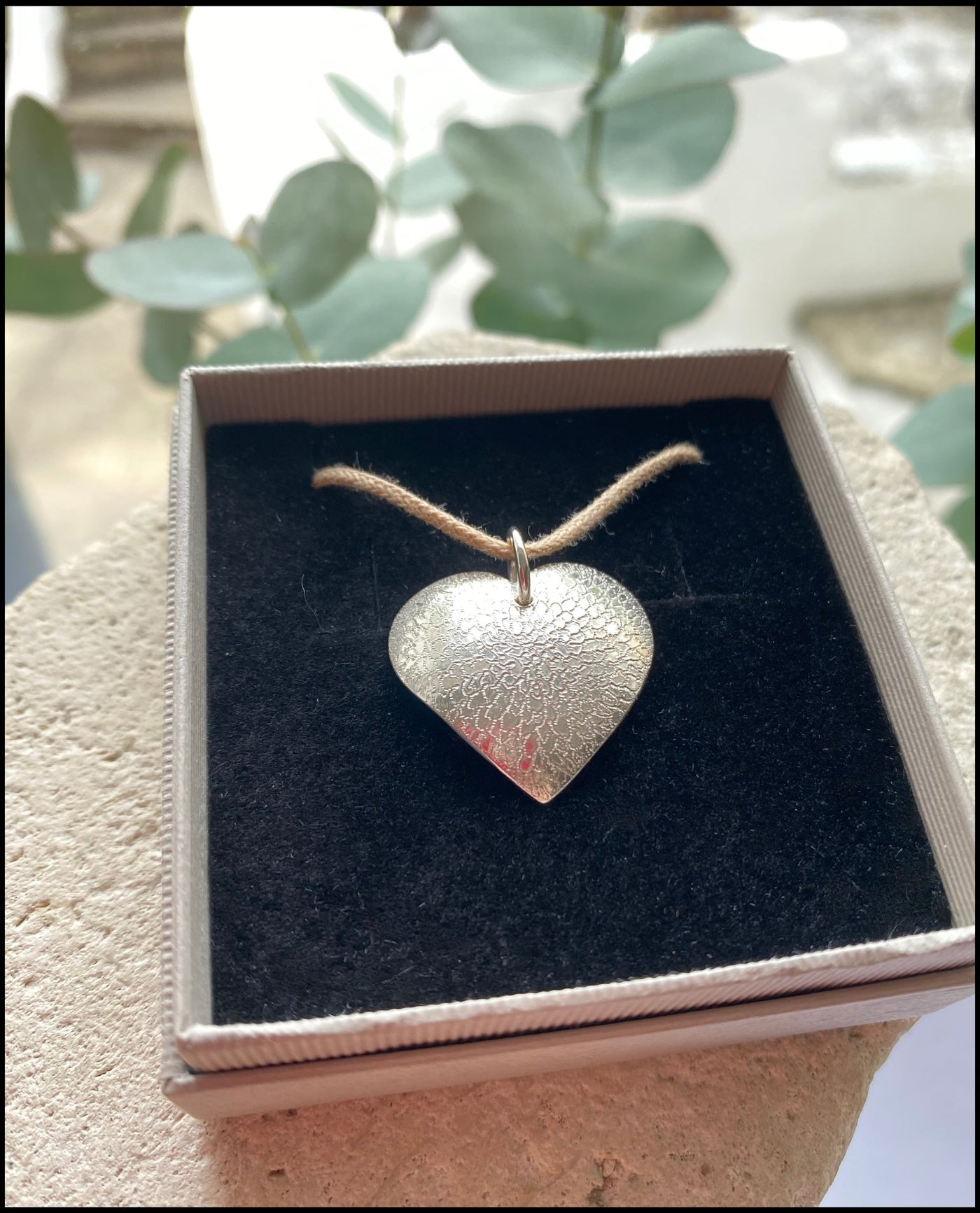 Silver heart-shaped pendant on a cotton cord in a box with greenery in the background