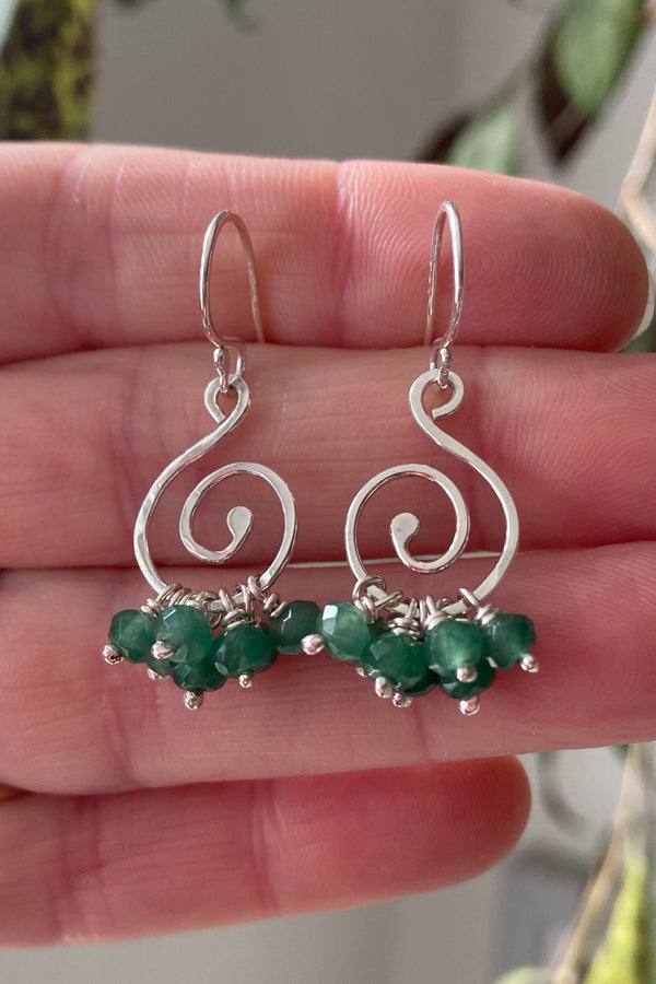 spiral earrings in silver with a ruffle of emerald gemstones hanging from the base of the spiral, held on the hand that made them