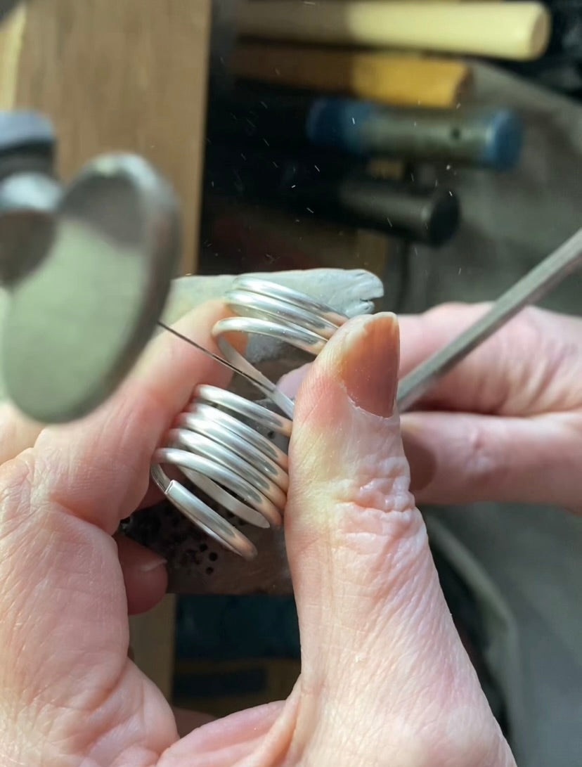 Close-up of hands forming a coiled silver ring using a saw frame in a workshop setting.
