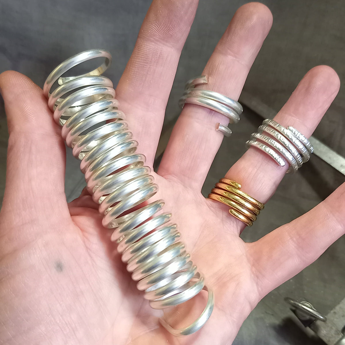 A hand holding a large coiled silver ring, with several smaller silver and gold rings displayed on the fingers, set against a dark gray textured background.
