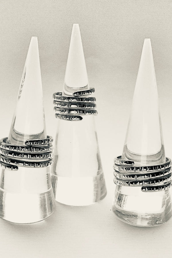 Three silver rings with textured, patterned bands displayed on clear cone-shaped ring stands against a light gray background.