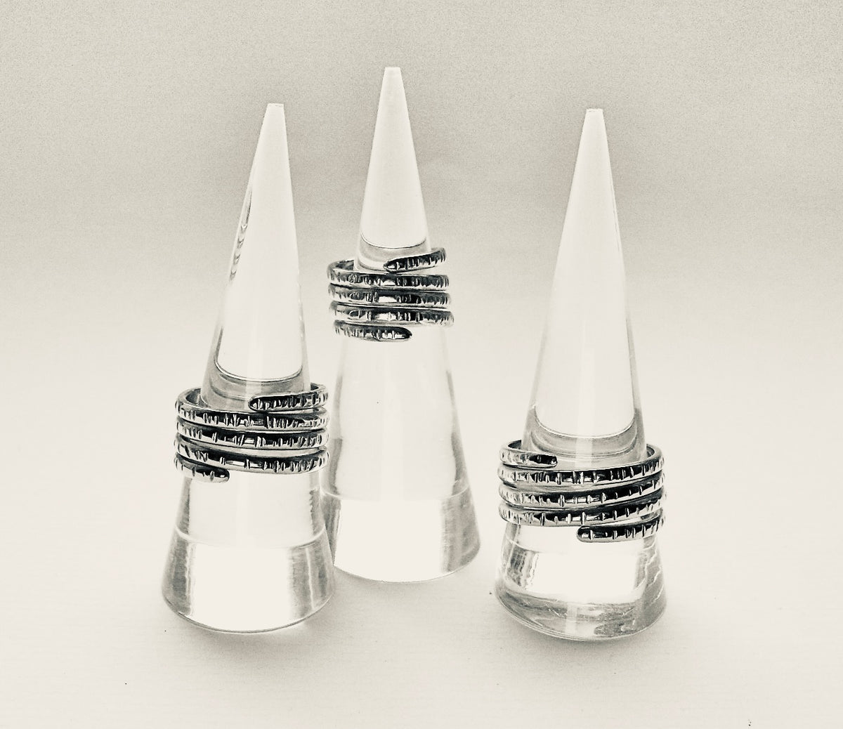 Three silver rings with textured, patterned bands displayed on clear cone-shaped ring stands against a light gray background.