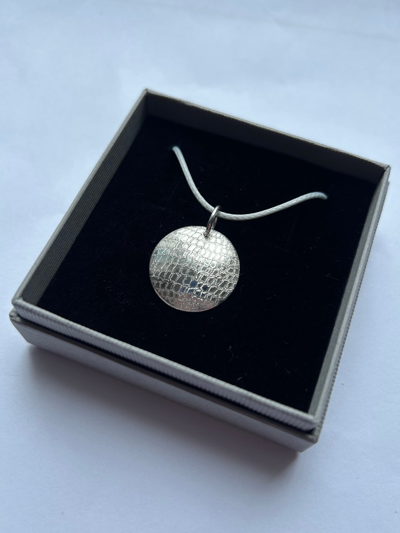 A silver pendant necklace with a textured, hand engraved finish representing a glitter ball -  resting inside a black-lined square gift box on a light gray background.