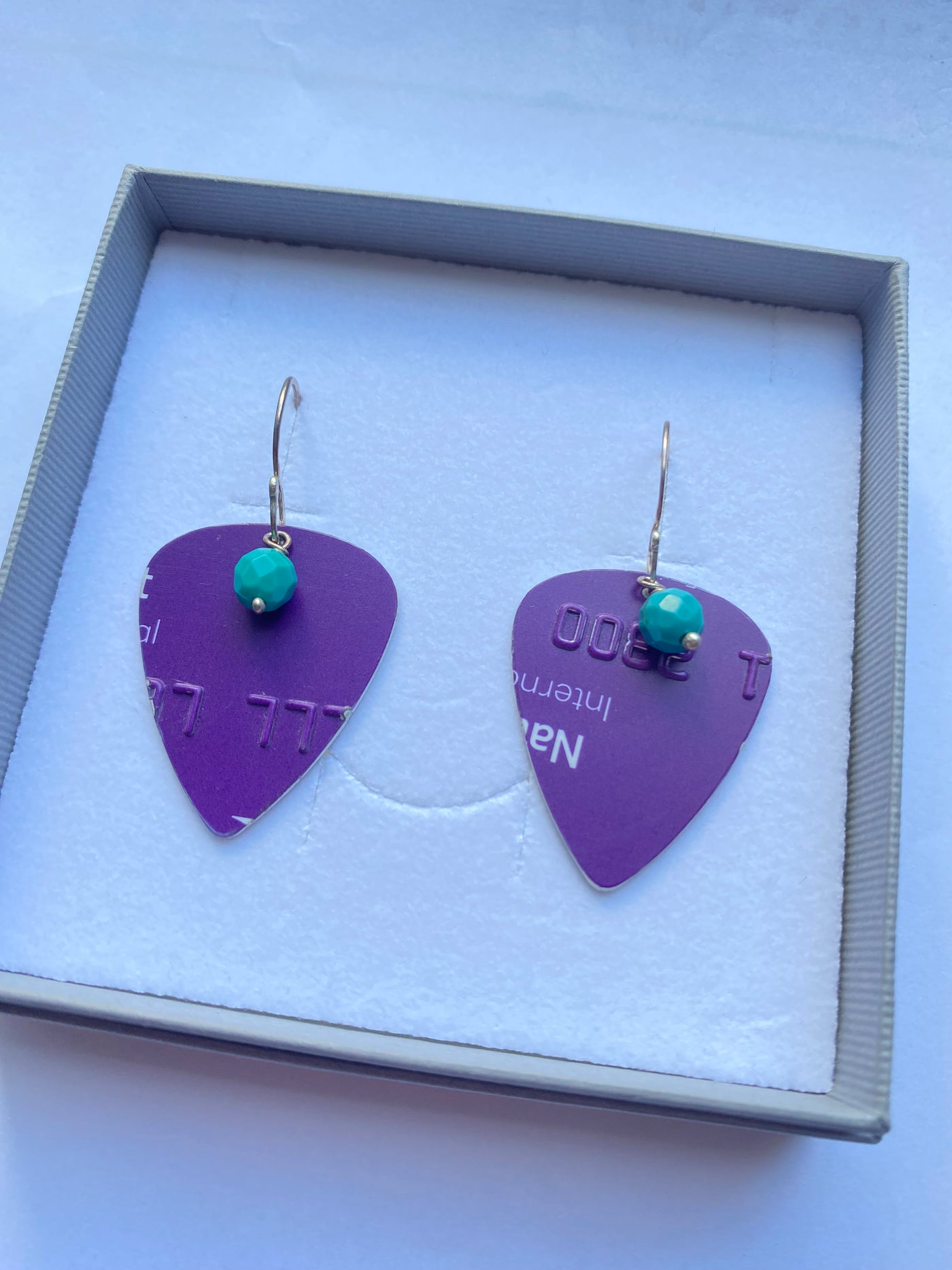 A pair of dangling earrings displayed in a gift box. Each earring has a purple guitar-pick-shaped drop with a small natural turquoise bead and a silver handmade fishhook ear wire.