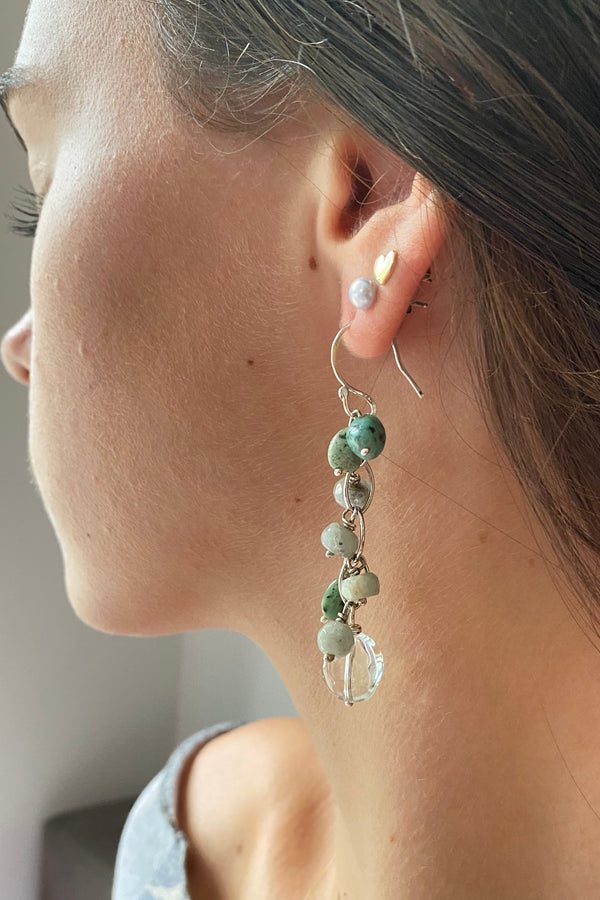 Close-up of an ear wearing a delicate earring with green beads on a neutral background. Beautiful waterfall earrings handmade in sterling silver with pale green prasiolite, dark green blue natural turquoise and sea foam coloured faceted aquamarine gemstones. 
