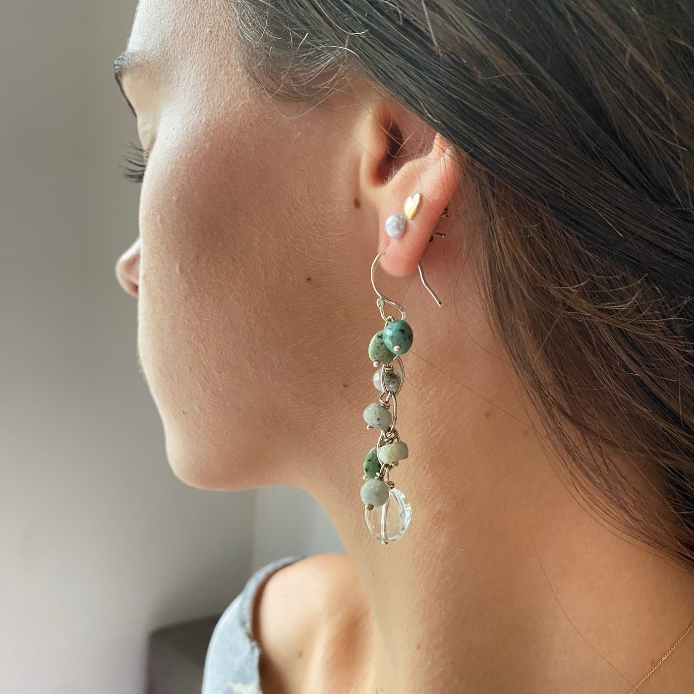 Close-up of an ear wearing a delicate earring with green beads on a neutral background. Beautiful waterfall earrings handmade in sterling silver with pale green prasiolite, dark green blue natural turquoise and sea foam coloured faceted aquamarine gemstones. 