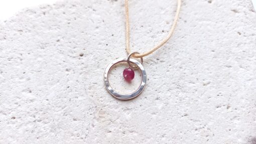 small textured silver circle pendant with small ruby dangle stone in the centre lying on stonel