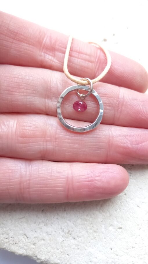 Textured silver open circle with little ruby in the centre hanging over a hand